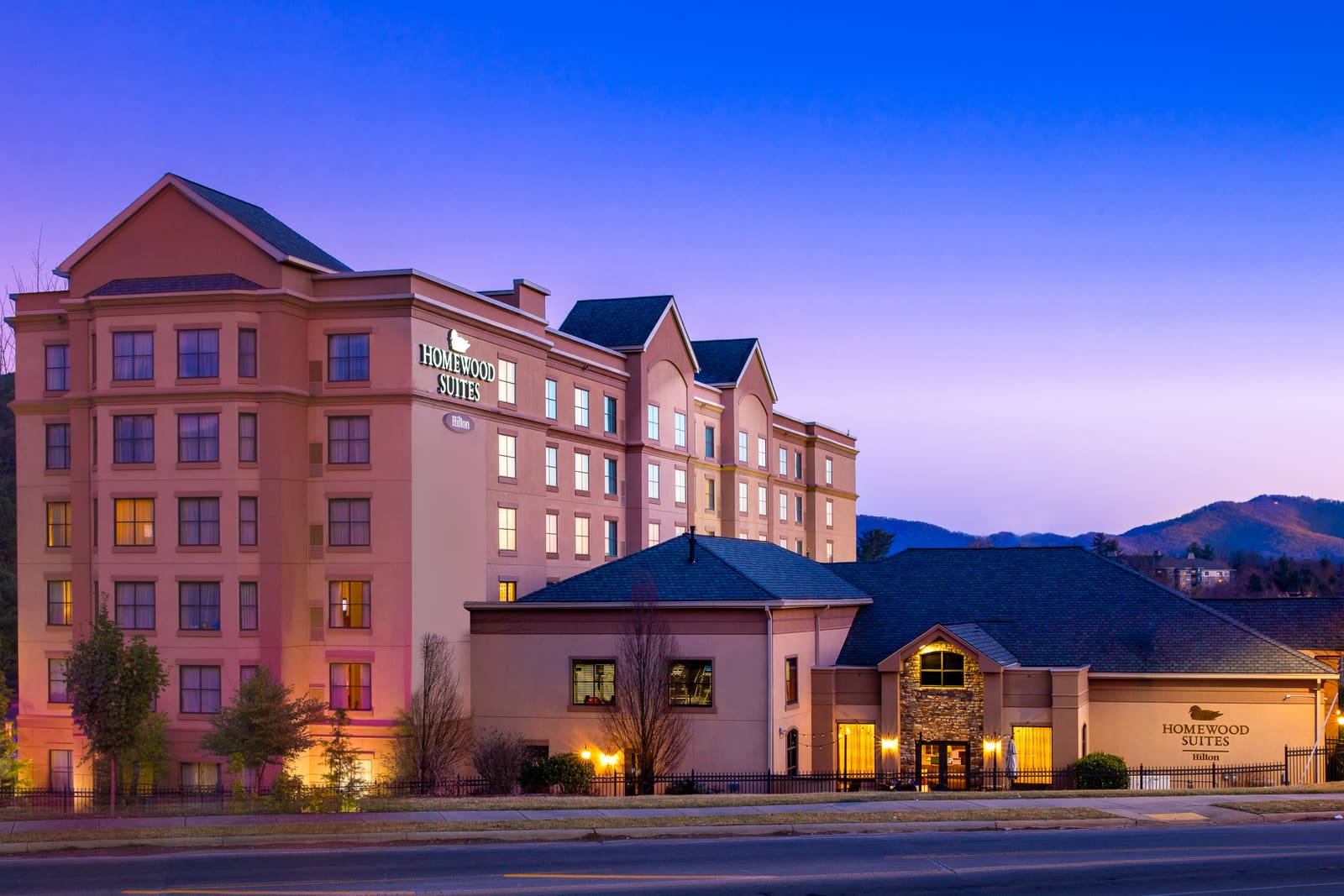 Homewood Suites by Hilton Asheville lit at dusk, with a mountain ridgeline rising behind the building.